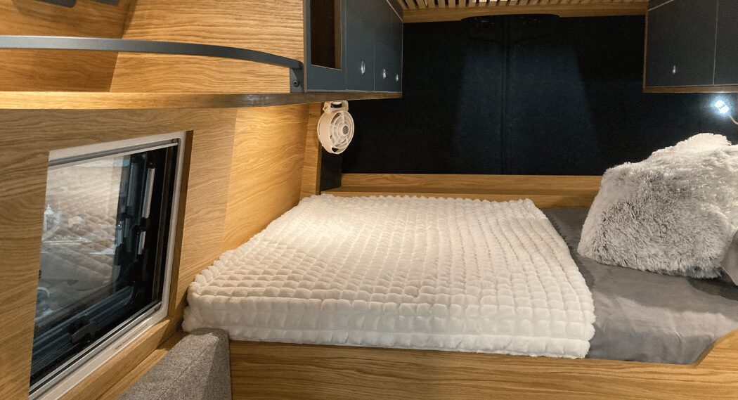 Intérieur d'un camping-car avec lit, coussins, bois clair et fenêtre à gauche.