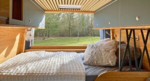 Intérieur de camping-car avec lit douillet, coussins, armoires et vue sur un paysage boisé.