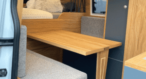 Intérieur d'un camping-car avec sièges gris, table en bois et lit à l'arrière.