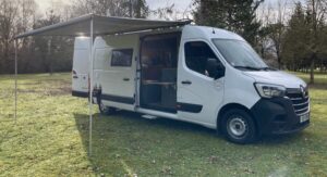 Fourgonnette blanche camping-car avec auvent, sur pelouse, arbres en arrière-plan.