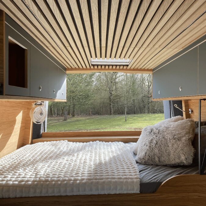 Intérieur van aménagé, lit, rangements gris, plafond bois, fenêtre vue sur un paysage arboré.