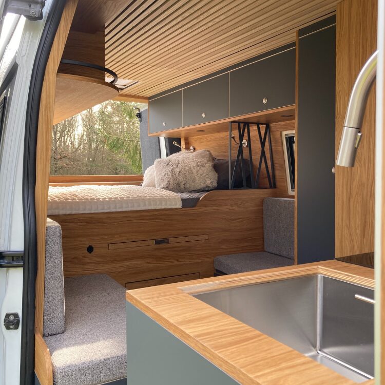 Intérieur de van aménagé en bois clair avec lit, rangements et lavabo en acier inoxydable.