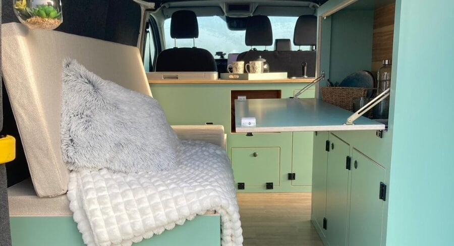 Intérieur d’un van aménagé avec canapé, couverture, coussin, placards et table rabattable.