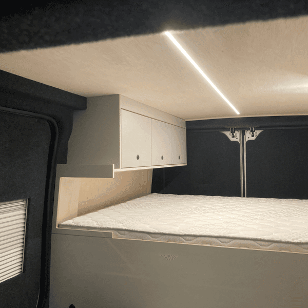 Intérieur d'un fourgon aménagé avec lit, armoires au-dessus et éclairage LED au plafond.
