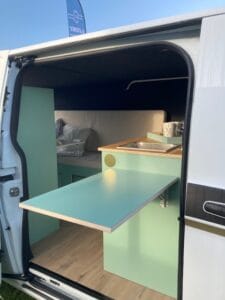 Camionnette aménagée avec lit et coin cuisine équipé d'un évier et d'une table pliable.