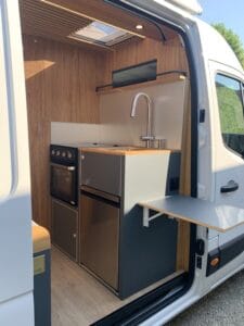 Intérieur d'un fourgon aménagé avec cuisine compacte, évier, four et table pliante en bois clair.