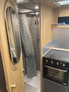 Petite salle de bain avec douche, miroir ovale, cuisine équipée avec four et plaque de cuisson.