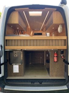 Intérieur de fourgon aménagé avec lit surélevé, bois, éclairage intégré, rangement, extincteur.