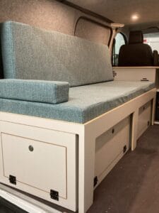 Banquette avec coussins bleus dans un véhicule aménagé, rangement dessous avec portes à charnières.