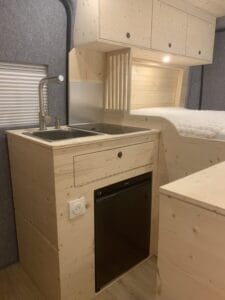 Cuisine en bois clair avec évier double, robinet, réfrigérateur noir, placards et prise.