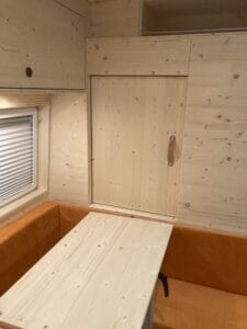 Intérieur bois clair avec table fixée au mur, sièges orange, fenêtre à store à gauche.