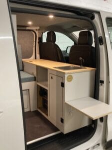 Vue intérieure d'un van aménagé avec kitchenette, évier, rangements, et plan de travail rabattable.