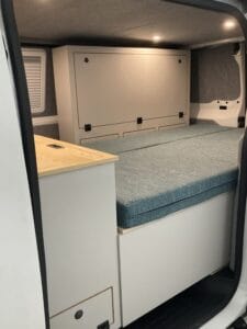 Aménagement de van avec lit, rangements, éclairage au plafond et surface de travail en bois.