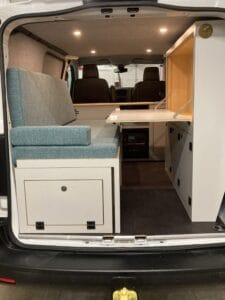 Aménagement intérieur de van avec sièges bleus, bureau rabattable et rangements, éclairage encastré.