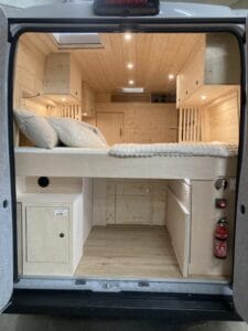 Intérieur d'un van aménagé en bois clair avec lit, rangements, éclairage et extincteur proche.