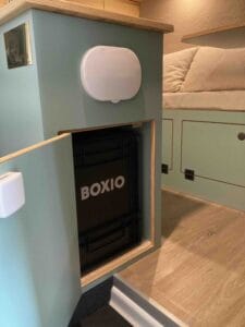 Boîte noire "Boxio" dans compartiment ouvert près d'un lit, sur sol en bois clair.