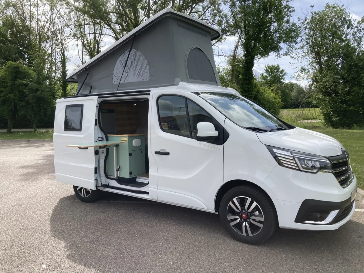 Camionnette blanche aménagée en camping avec toit relevé, porte ouverte, en extérieur verdoyant.