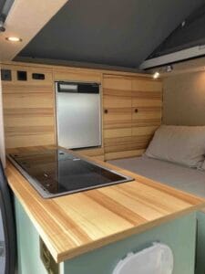 Intérieur de camionnette aménagée avec lit, comptoir bois, plaque cuisson, placards et réfrigérateur.